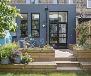 A raised deck outside a new kitchen extension with outdoor bistro style seating and raised beds made from decking planks and steps down to a lawn area